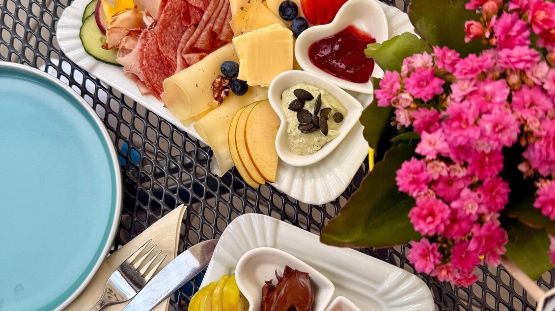 Ein appetitliches Frühstück mit Wurst, Käse, frischen Früchten und verschiedenen Dips. Die Teller sind auf einem dekorativen Tisch mit bunten Blumen angerichtet. | © Lisa-Marie Schiefer