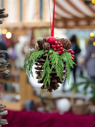Ein dekorativer Tannenzapfen mit grünem Blattwerk und roten Beeren hängt in einem festlichen Umfeld. Im Hintergrund sind verschwommene Lichter und Stände eines Weihnachtsmarkts zu sehen. | © Pixabay