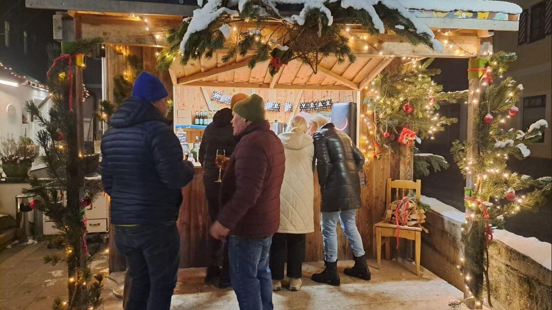 Ein festlich dekorierter Stand mit Weihnachtsbeleuchtung in einer schneebedeckten Umgebung. Menschen stehen zusammen und genießen Getränke in gemütlicher Atmosphäre. | © Bio Hotel Tiefenbach