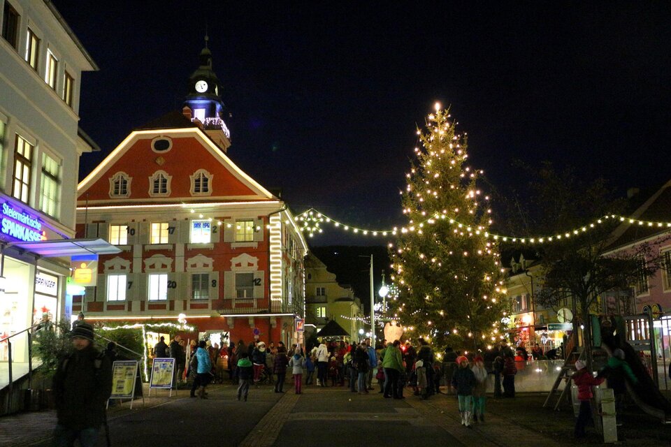 Eine festliche Stadtansicht bei Nacht mit einem beleuchteten Weihnachtsbaum. Es gibt viele Menschen auf der Straße, die die festliche Atmosphäre genießen.