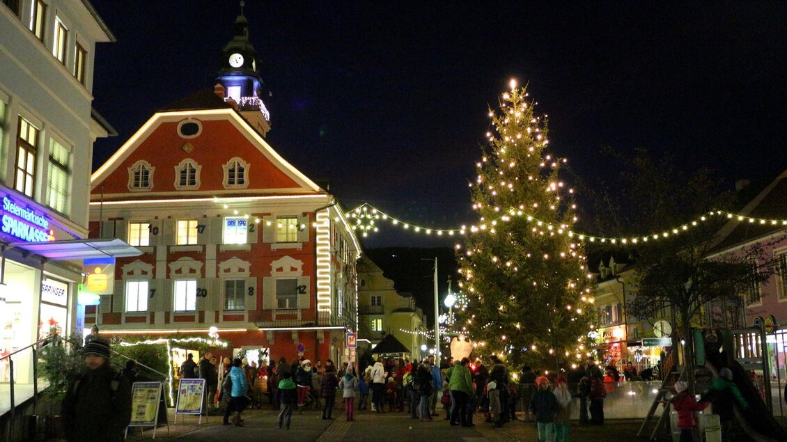 Eine festliche Stadtansicht bei Nacht mit einem beleuchteten Weihnachtsbaum. Es gibt viele Menschen auf der Straße, die die festliche Atmosphäre genießen.