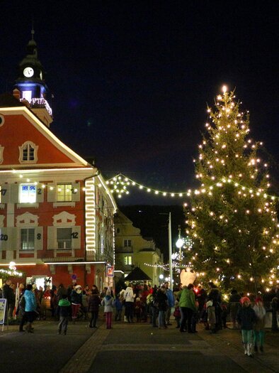 Eine festliche Stadtansicht bei Nacht mit einem beleuchteten Weihnachtsbaum. Es gibt viele Menschen auf der Straße, die die festliche Atmosphäre genießen.