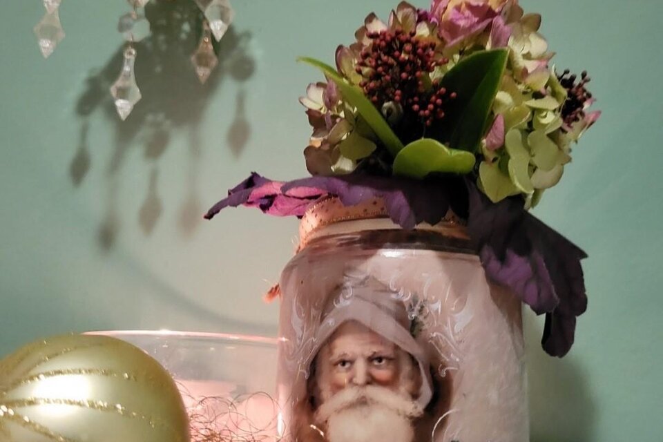 Ein dekoriertes Glas mit einem Bild von einem alten Mann und bunten Blumen. Daneben liegen goldene Dekorationen und eine glitzernde Kugel. | © Malermeisterbetrieb Enzi