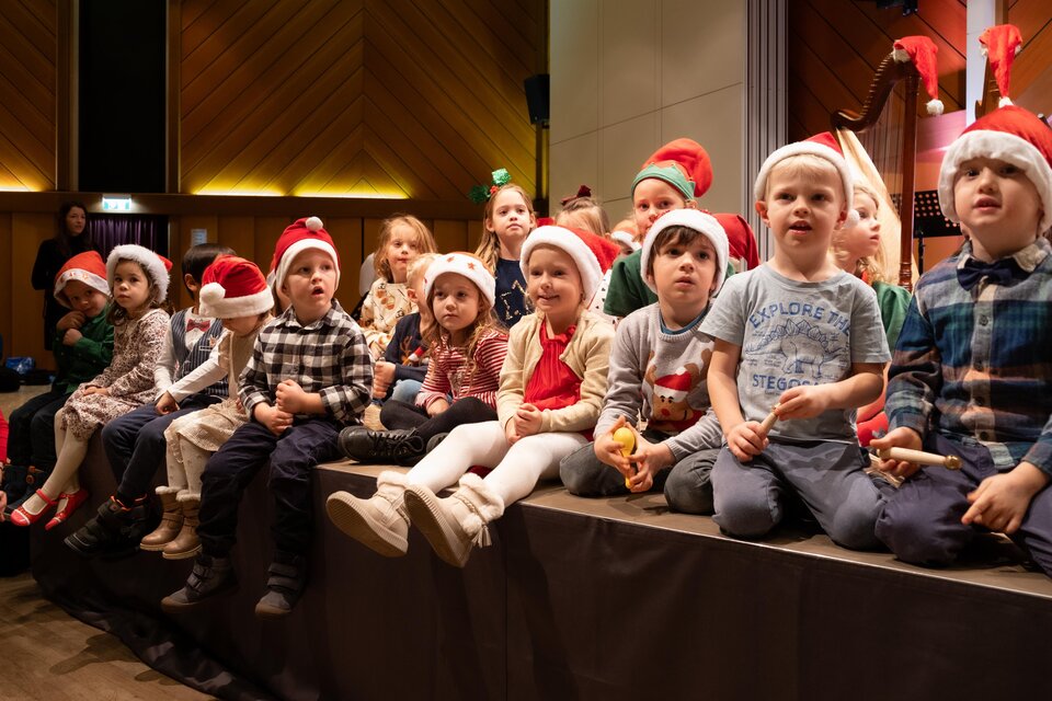 Eine Gruppe von Kindern sitzt auf einer Bühne, alle tragen Weihnachtsmützen. Sie schauen gespannt und lächeln, während sie an einem weihnachtlichen Event teilnehmen. | © Petra Polácková