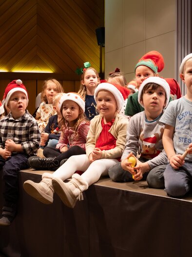 Eine Gruppe von Kindern sitzt auf einer Bühne, alle tragen Weihnachtsmützen. Sie schauen gespannt und lächeln, während sie an einem weihnachtlichen Event teilnehmen. | © Petra Polácková