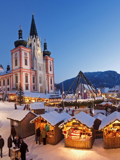 A festive Christmas market with wooden huts and many visitors in a snowy town. In the background, there is a historical building under a clear evening sky. | © Fred Lindmoser