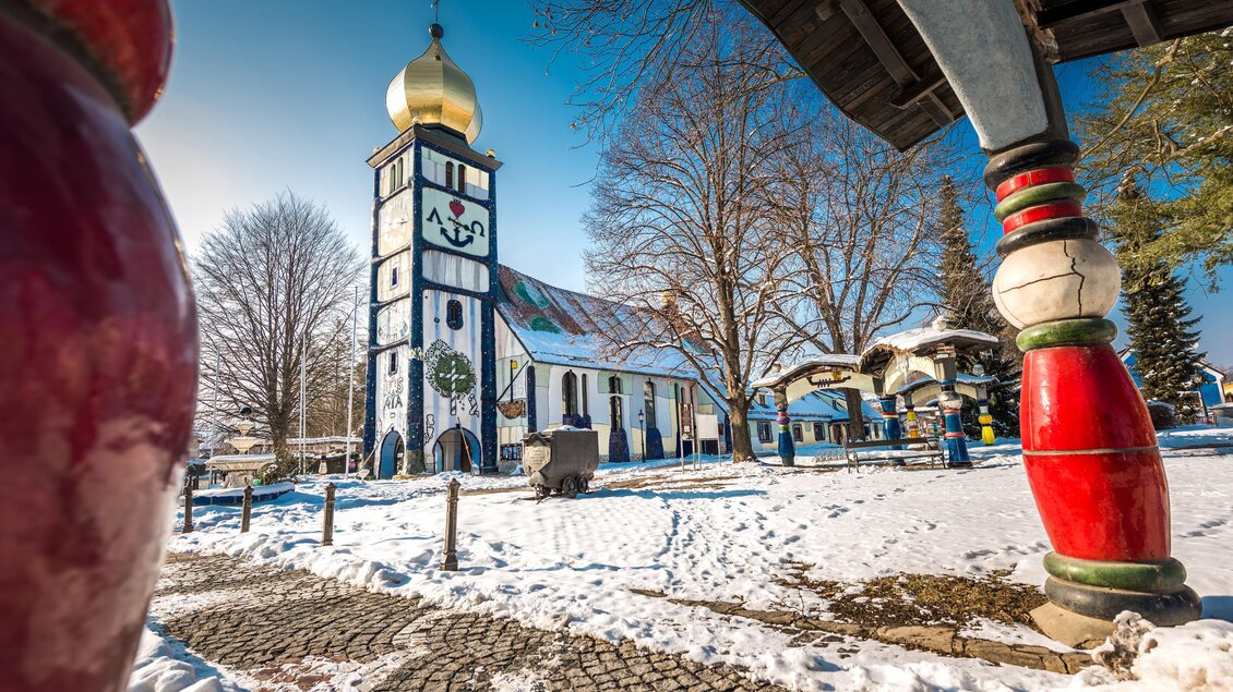 Eine bunte Kirche mit einer goldenen Kuppel steht in der Winterlandschaft. Der Boden ist von Schnee bedeckt und es sind einige Bäume im Hintergrund zu sehen. | © Lipizzanerheimat-DieAbbilderei