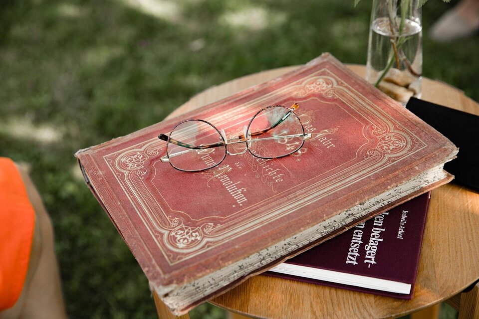 Ein alter, roter Buchdeckel liegt auf einem Tisch, darauf liegen eine Brille und ein weiteres Buch. Im Hintergrund sind grüne Wiesen zu sehen. | © TV Südsteiermark - achromaticphotography
