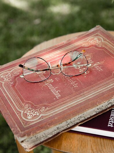 An old red book cover lies on a table, on top of it are a pair of glasses and another book. In the background, green meadows can be seen. | © TV Südsteiermark - achromaticphotography