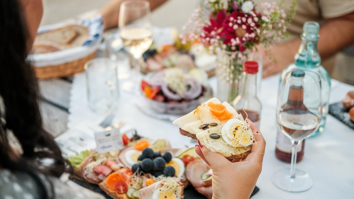 Ein Tisch gedeckt mit köstlichem Essen, darunter belegte Brote und frisches Gemüse. Im Hintergrund sieht man Gläser mit Wasser und Wein sowie einen Blumenstrauß. | © MALU