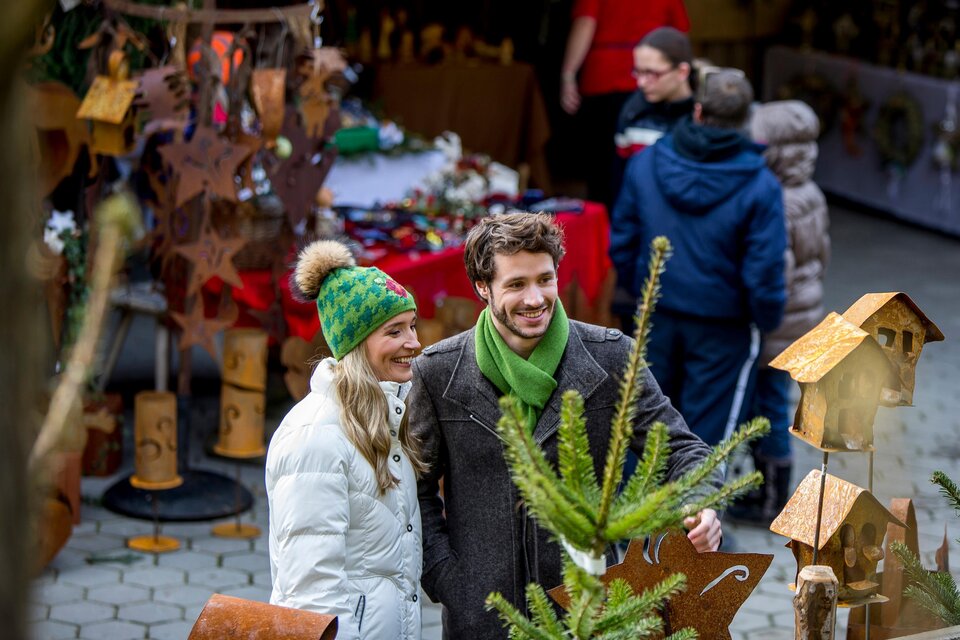 Ein Paar lächelt in einem festlichen Weihnachtsmarkt, umgeben von handgemachten Ornamenten und Dekorationen. Im Hintergrund sind weitere Besucher zu sehen, die den Markt erkunden. | © Region Graz-Tom Lamm