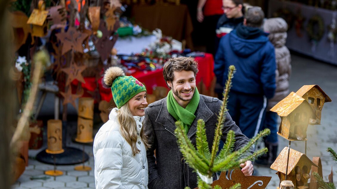Ein Paar lächelt in einem festlichen Weihnachtsmarkt, umgeben von handgemachten Ornamenten und Dekorationen. Im Hintergrund sind weitere Besucher zu sehen, die den Markt erkunden. | © Region Graz-Tom Lamm