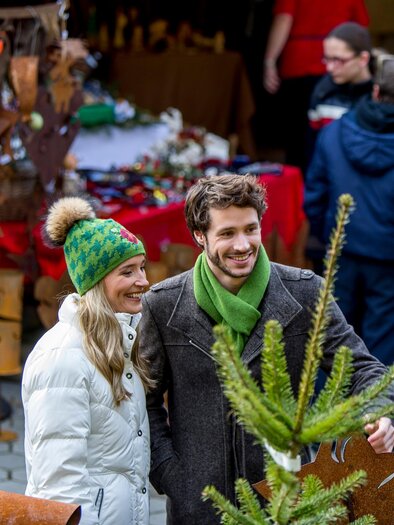 A couple smiles at a festive Christmas market, surrounded by handmade ornaments and decorations. In the background, other visitors can be seen exploring the market. | © Region Graz-Tom Lamm