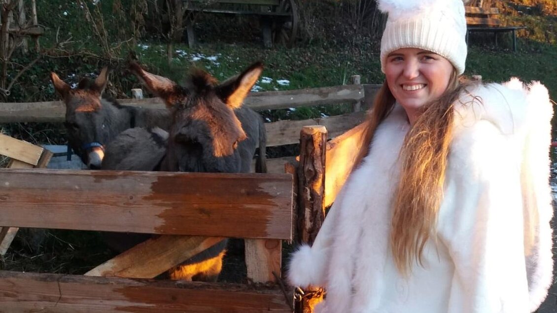 Eine Person in einem weißen Outfit steht neben einem Zaun und lächelt. Im Hintergrund sind zwei Esel zu sehen. | © Sylvia Loidolt