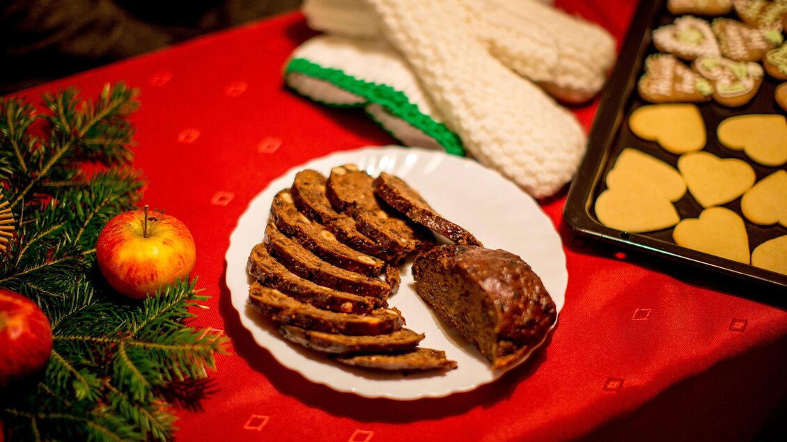 Ein festlich gedeckter Tisch mit einem Teller von geschnittenem Weihnachtsgebäck und frischen Äpfeln. Im Hintergrund sind Plätzchen in verschiedenen Formen auf einem Backblech zu sehen. | © Region Graz-Tom Lamm