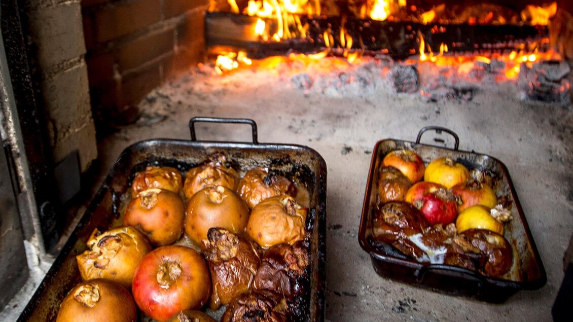 Ein traditioneller Ofen mit einem Feuer, in dem gebratene Äpfel auf zwei Blechträgern zubereitet werden. Die Äpfel sind unterschiedlich gefärbt und sehen köstlich aus. | © Region Graz-Tom Lamm