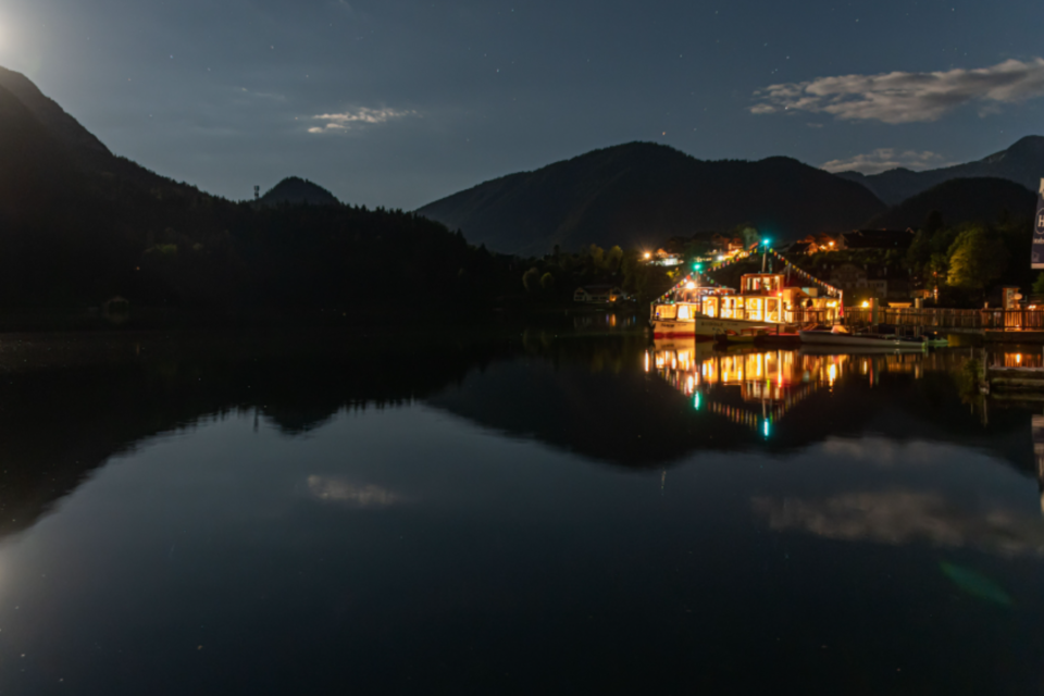 Ein ruhiger See bei Nacht, umgeben von Bergen und sanften Wolken. Das Licht des Mondes spiegelt sich auf der Wasseroberfläche. | © Schiffsfahrt Grundlsee