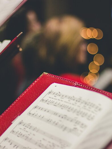 A choir holding musical notes in red folders. In the background, there are blurry lights and singing people. | © david-beale-unsplash