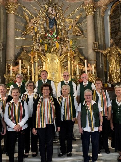 A group of singers stands in front of a magnificent altar in a church. They all wear festive clothing and a striped scarf. | © Vulkanlandchor Pertlstein
