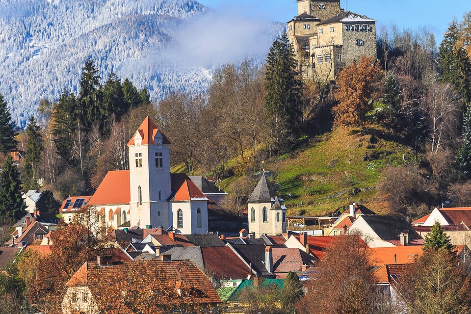 Eine malerische Stadt mit roten Dächern und einer Kirche im Vordergrund. Im Hintergrund steht eine alte Burg auf einem bewaldeten Hügel.