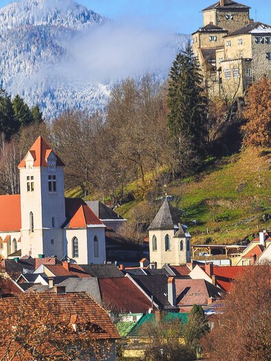 Eine malerische Stadt mit roten Dächern und einer Kirche im Vordergrund. Im Hintergrund steht eine alte Burg auf einem bewaldeten Hügel.
