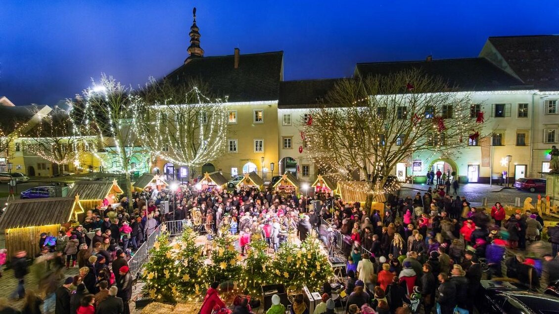 Ein belebter Weihnachtsmarkt mit vielen Menschen und festlicher Beleuchtung. Umgeben von traditionellen Ständen und geschmückten Bäumen strahlt die Atmosphäre Gemütlichkeit aus. | © TV Thermen- & Vulkanland