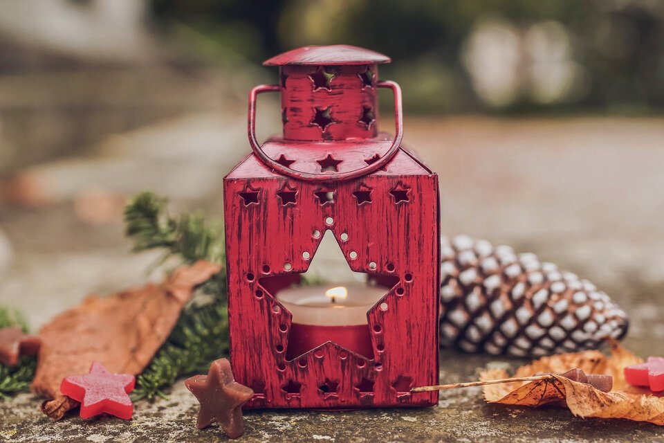 Eine rote Laterne mit einem Stern und einer Kerze steht auf dem Boden. Um sie herum liegen Blätter, Sterne und eine Tannenzapfen. | © pixabay