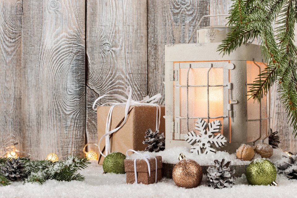 Eine rustikale Weihnachtsdekoration mit einer Laterne, Geschenken und Tannenzweigen. Die Szene ist auf weißem Kunstschnee und einer hölzernen Wand arrangiert. | © AdobeStock