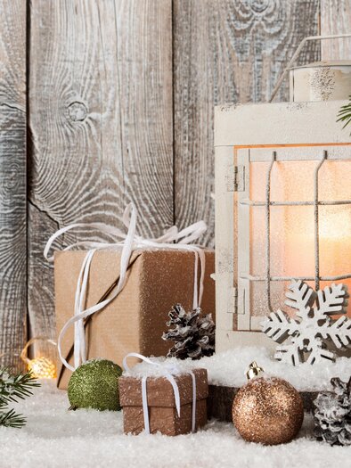 Eine rustikale Weihnachtsdekoration mit einer Laterne, Geschenken und Tannenzweigen. Die Szene ist auf weißem Kunstschnee und einer hölzernen Wand arrangiert. | © AdobeStock