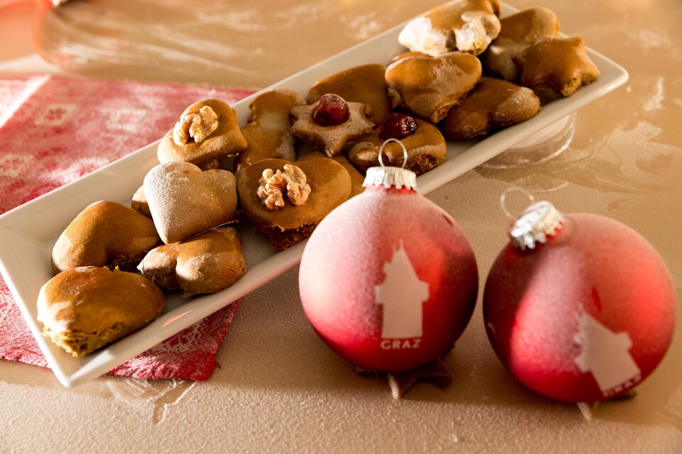Eine Platte mit verschiedenen Weihnachtsplätzchen liegt neben zwei dekorativen Weihnachtskugeln. Die Plätzchen sind in unterschiedlichen Formen und dekoriert, während die Kugeln rot und auffällig sind. | © Region Graz-Harry Schiffer