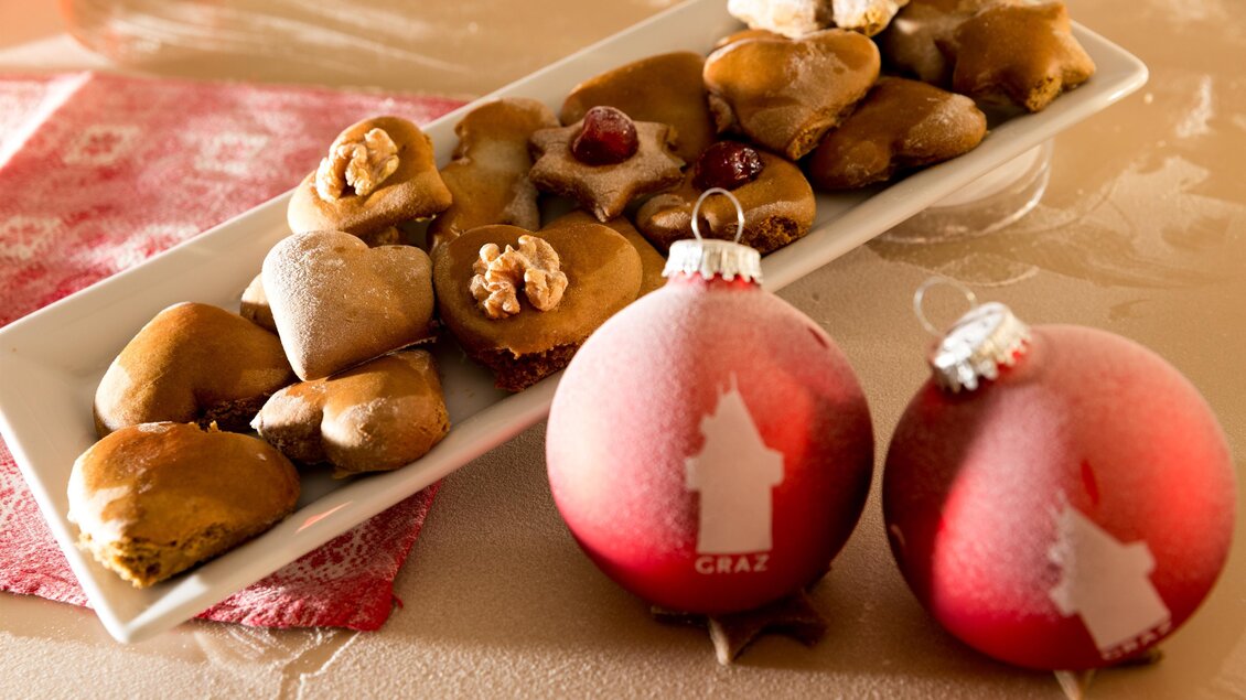 Eine Platte mit verschiedenen Weihnachtsplätzchen liegt neben zwei dekorativen Weihnachtskugeln. Die Plätzchen sind in unterschiedlichen Formen und dekoriert, während die Kugeln rot und auffällig sind. | © Region Graz-Harry Schiffer