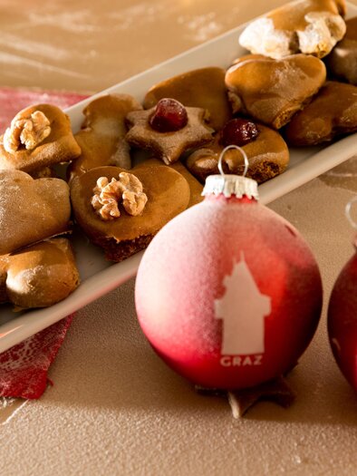 A plate with various Christmas cookies lies next to two decorative Christmas ornaments. The cookies are in different shapes and decorated, while the ornaments are red and striking. | © Region Graz-Harry Schiffer