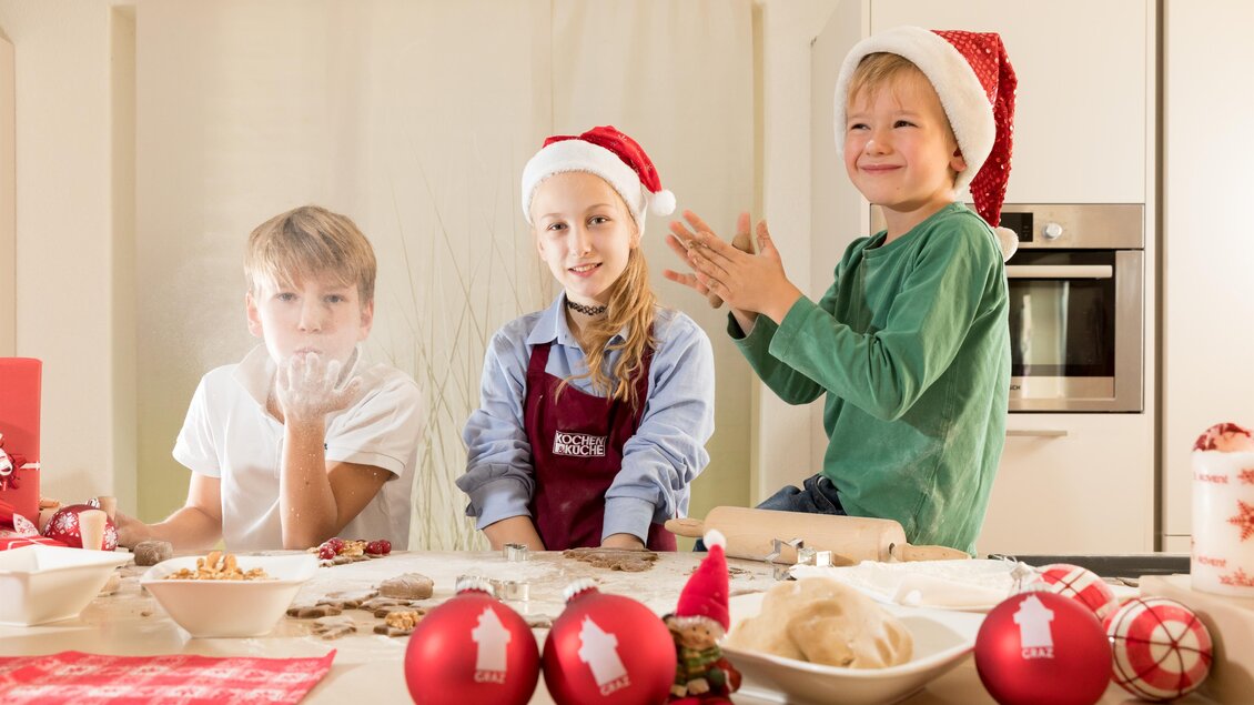 Drei Kinder mit Weihnachtsmützen stehen in einer Küche und haben Spaß beim Backen. Auf dem Tisch sind Zutaten und Dekoration für festliche Leckereien zu sehen. | © Region Graz-Harry Schiffer