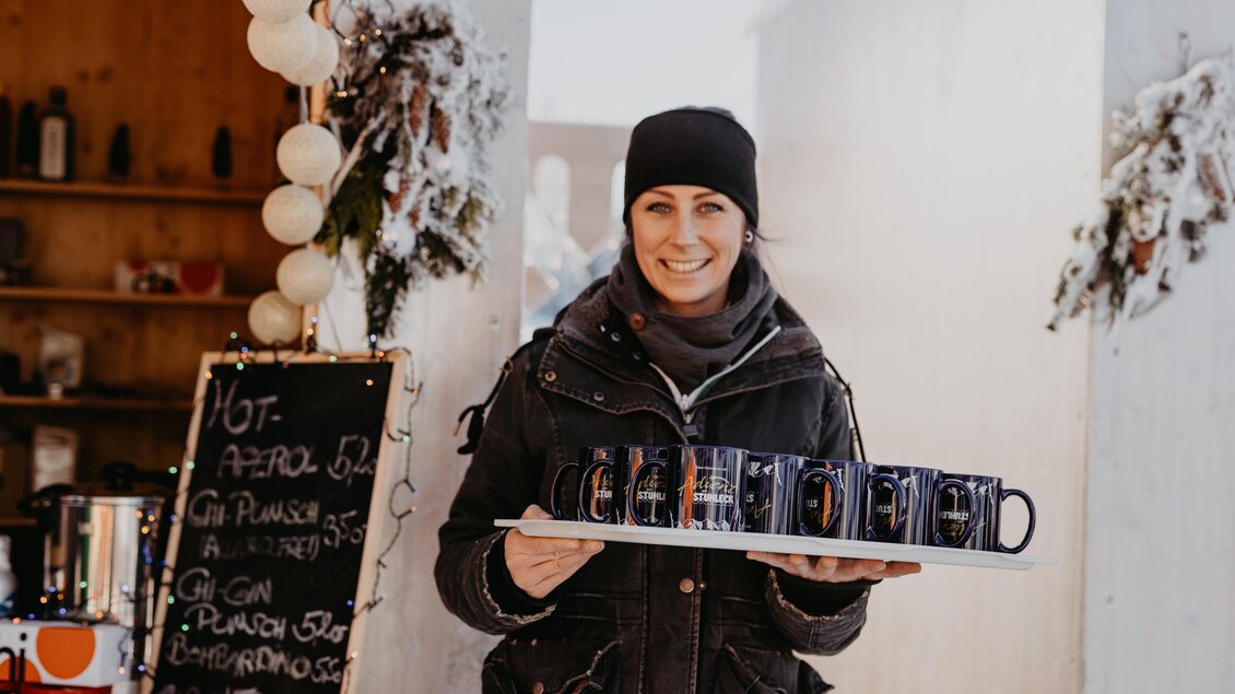 Eine lächelnde Frau trägt ein Tablett mit Getränken. Im Hintergrund sind eine Tafel mit Angeboten und festliche Dekoration zu sehen. | © nicoleseiser.at