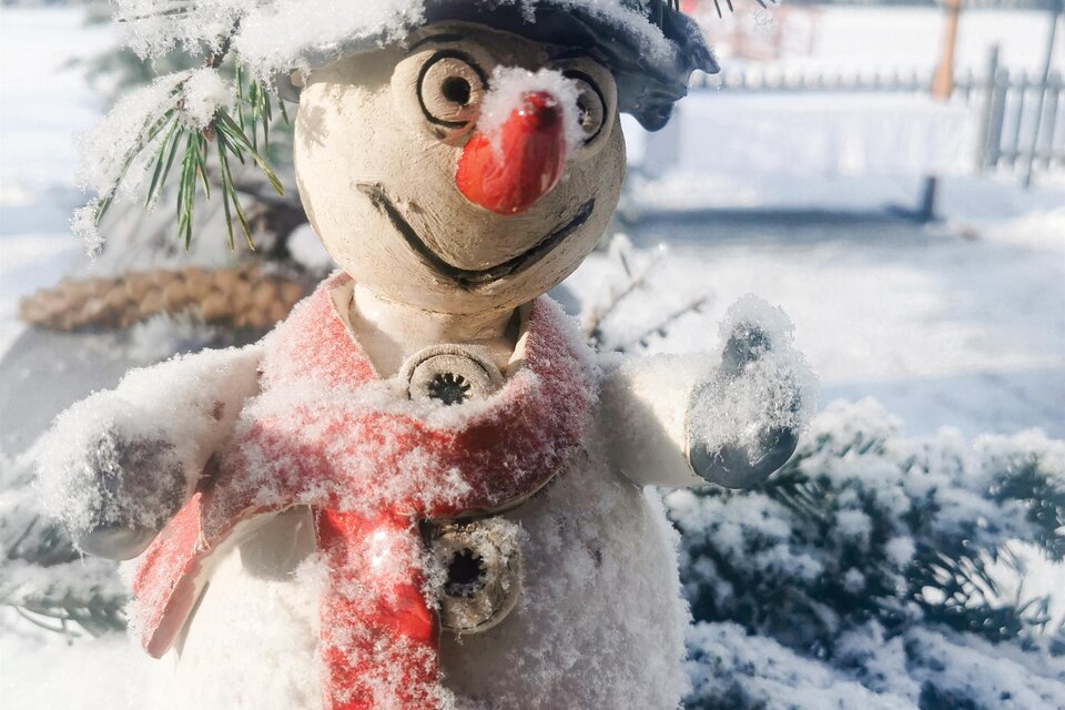 Ein fröhlicher Schneemann mit Karottennase und rotem Schal steht im Schnee. Er hat einen kleinen Hut und ist von verschneiten Tannen umgeben. | © Schuster Kulinarik