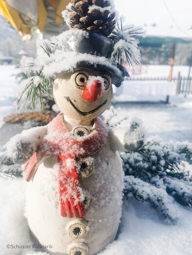 Ein fröhlicher Schneemann mit Karottennase und rotem Schal steht im Schnee. Er hat einen kleinen Hut und ist von verschneiten Tannen umgeben. | © Schuster Kulinarik