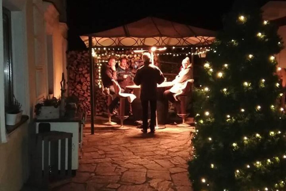 Eine gemütliche Terrasse bei Nacht mit einem Pavillon und Lichtern. Mehrere Personen sitzen zusammen und genießen die Atmosphäre.