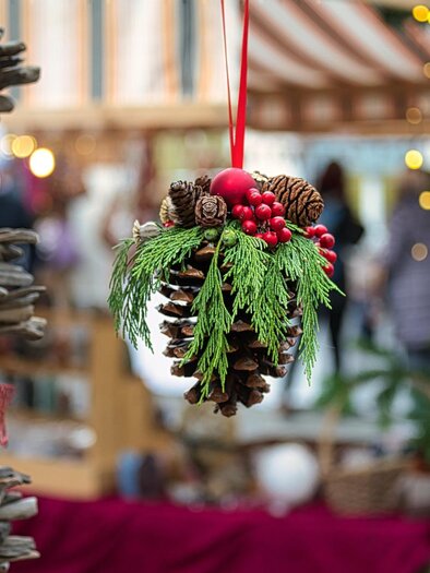 A beautiful Christmas decoration made from a pine cone base, surrounded by green branches and red berries. In the background, blurred people and lights can be seen. | © Pixabay