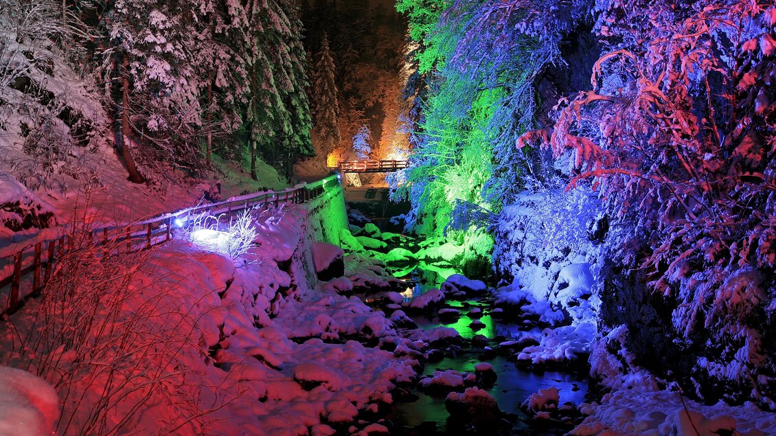 Eine verschneite Landschaft bei Nacht mit bunten Lichtern. Bäume und ein Bach sind in leuchtenden Farben beleuchtet. | © Herbert Raffalt