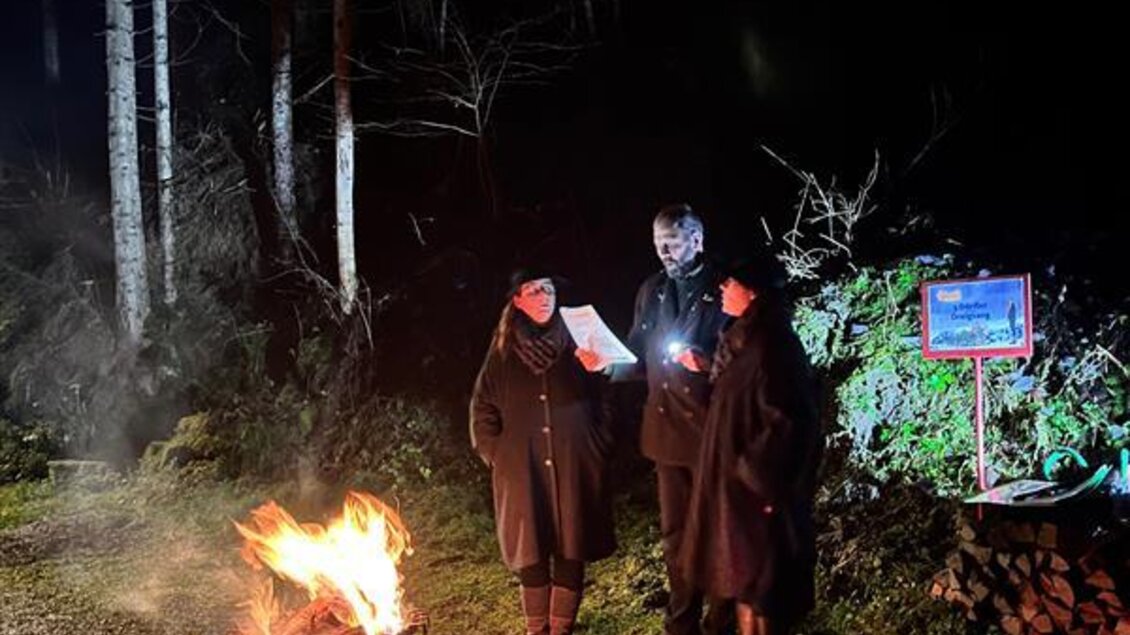 Drei Personen stehen nachts um ein Feuer in einem Korb. Im Hintergrund sind Bäume und ein Schild sichtbar. | © Herbert Raffalt