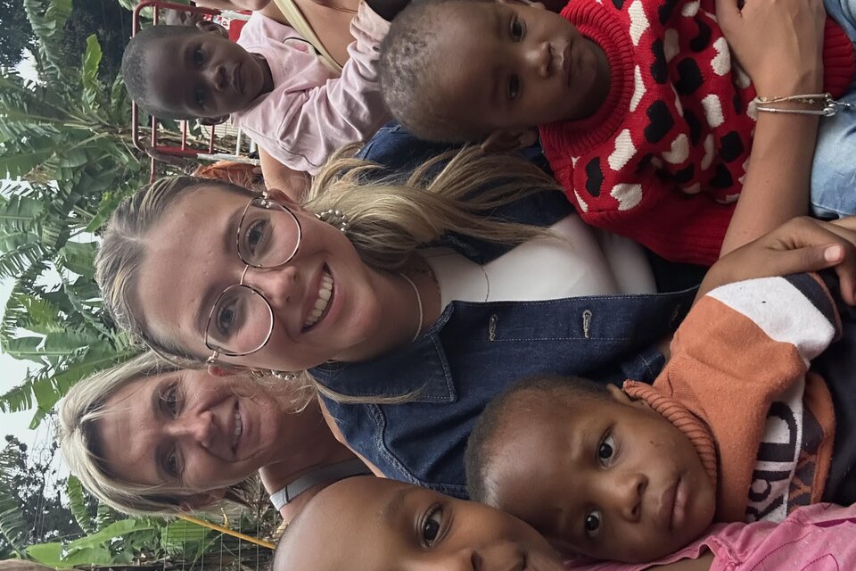 Gruppen-Selfie im Freien: zwei junge Frauen und eine ältere Frau lächeln in die Kamera, umgeben von mehreren Kindern; im Hintergrund ein Garten/Spielbereich mit Bäumen und Bananenstauden. | © Paula Daum