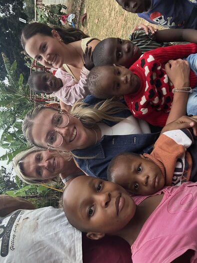 Gruppen-Selfie im Freien: zwei junge Frauen und eine ältere Frau lächeln in die Kamera, umgeben von mehreren Kindern; im Hintergrund ein Garten/Spielbereich mit Bäumen und Bananenstauden. | © Paula Daum