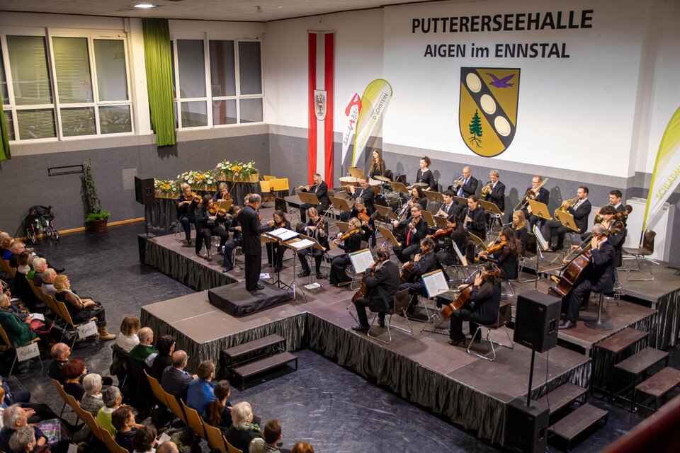 Ein Musikerlehrer leitet ein Orchester in der Putterssehalle. Das Publikum sitzt gebannt und genießt die Darbietung. | © Gemeinde Aigen im Ennstal