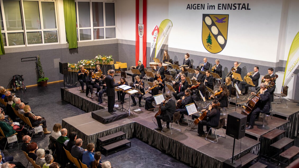 Ein Musikerlehrer leitet ein Orchester in der Putterssehalle. Das Publikum sitzt gebannt und genießt die Darbietung. | © Gemeinde Aigen im Ennstal
