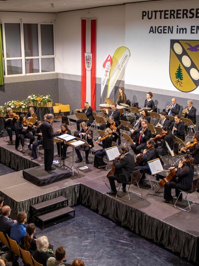 Ein Musikerlehrer leitet ein Orchester in der Putterssehalle. Das Publikum sitzt gebannt und genießt die Darbietung. | © Gemeinde Aigen im Ennstal