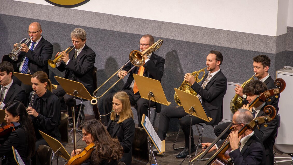Ein Orchester spielt auf einer Bühne. In der ersten Reihe sind Blasinstrumente und in der zweiten Reihe Streichinstrumente zu sehen. | © Gemeinde Aigen im Ennstal