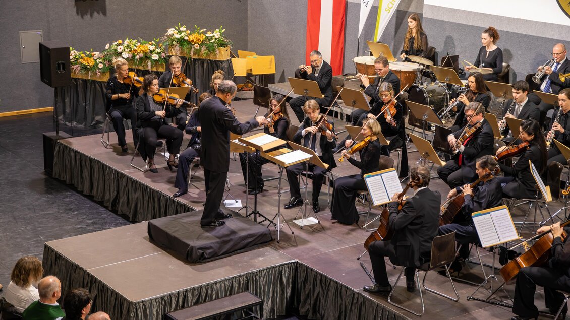 Ein Orchester spielt auf einer Bühne, während ein Dirigent die Musiker leitet. Im Hintergrund sind die Zuschauer und Blumenarrangements zu sehen. | © Gemeinde Aigen im Ennstal