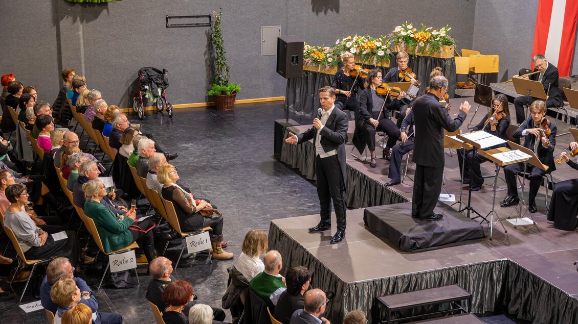 Ein Konzert in einem Saal mit einem Sänger und einem Orchester. Die Zuschauer sitzen aufmerksam im Publikum und lauschen der Darbietung. | © Gemeinde Aigen im Ennstal
