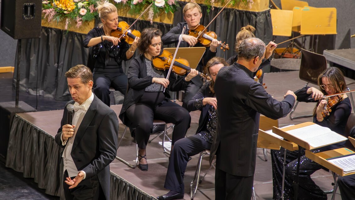 Ein Musiker steht im Vordergrund und singt, während ein Orchester im Hintergrund spielt. Die Instrumentalisten sind elegant gekleidet und konzentriert bei der Aufführung. | © Gemeinde Aigen im Ennstal