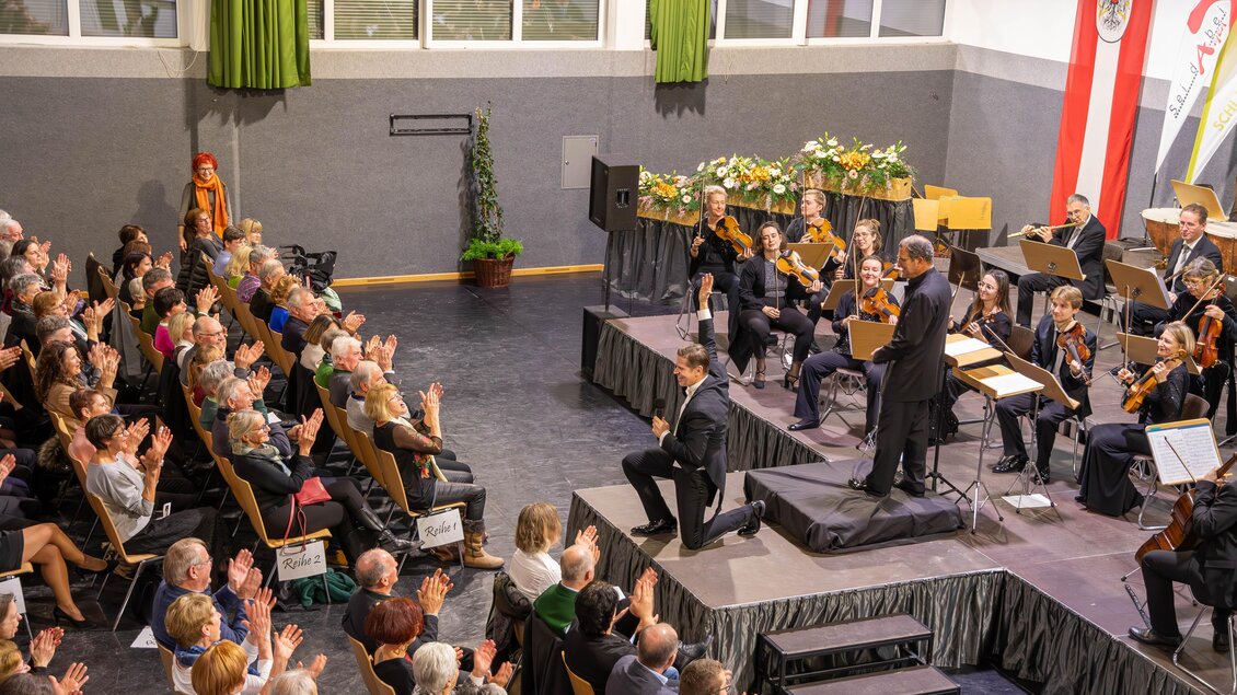 Ein Konzertsaal voller begeisterter Zuschauer applaudiert. Auf der Bühne steht ein Dirigent vor einem Orchester. | © Gemeinde Aigen im Ennstal